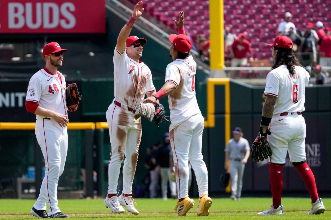 Four Cincinnati Reds baseball players in white uniforms and red caps exchange high fives on the field.